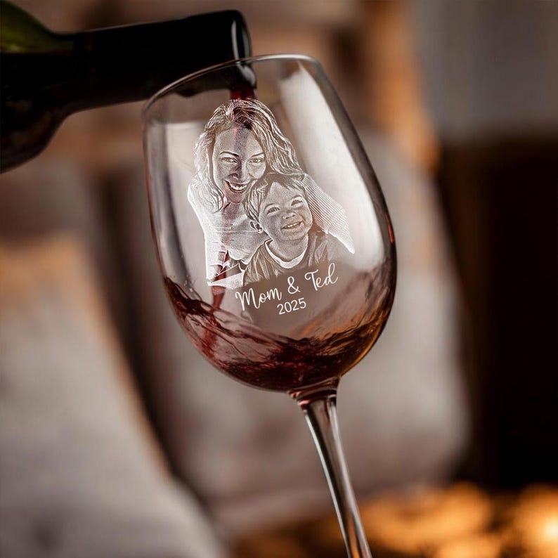 Pode incluir: Uma ta&ccedil;a de vinho transparente com um design gravado personalizado de uma m&atilde;e e um filho, com o texto "Mom & Ted 2025". O vinho tinto est&aacute; sendo derramado na ta&ccedil;a de uma garrafa escura. A ta&ccedil;a tem uma haste longa.