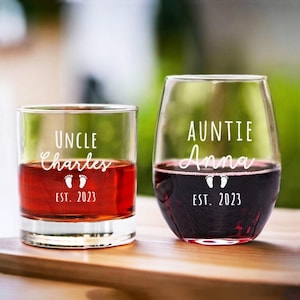 May include: Two personalized glasses, one a rocks glass and the other a stemless wine glass, filled with red liquid. The rocks glass reads "UNCLE Charles" with footprints and "EST. 2023". The wine glass reads "AUNTIE Anna" with footprints and "EST. 2023".
