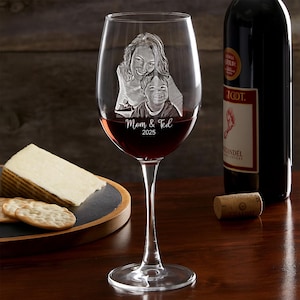 Pode incluir: Um copo de vinho transparente cheio de vinho tinto, com uma imagem gravada de uma m&atilde;e e uma crian&ccedil;a com o texto "Mom & Ted 2025". Uma garrafa de vinho tinto e um prato de queijo e bolachas est&atilde;o no fundo.