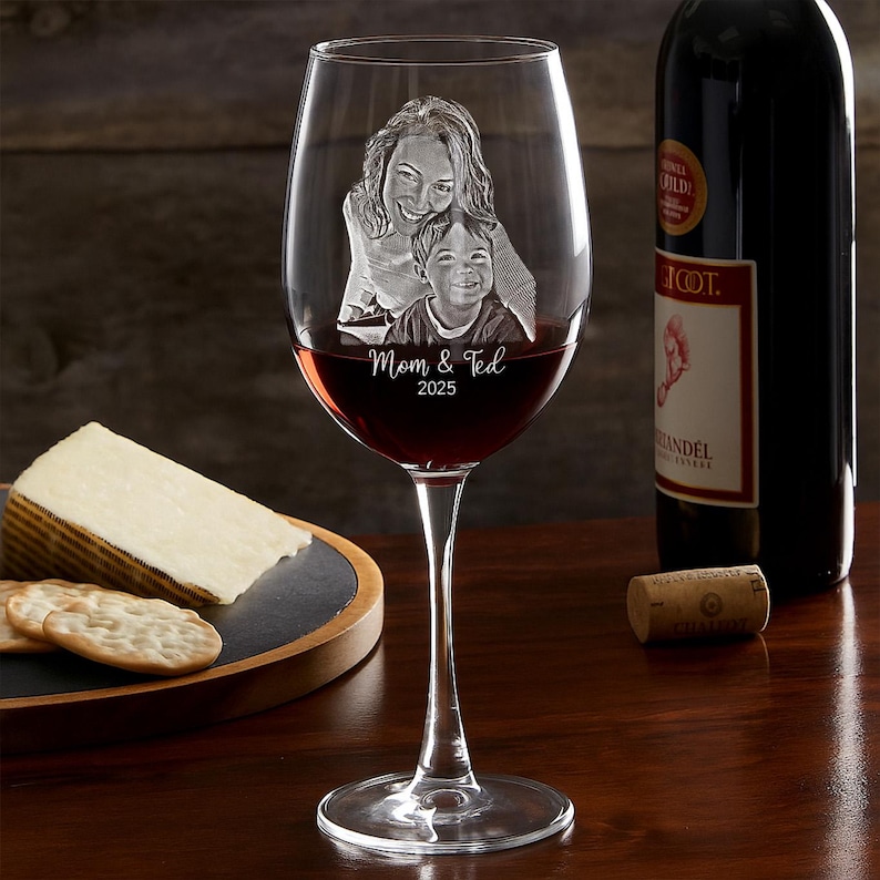 Pode incluir: Um copo de vinho transparente cheio de vinho tinto, com uma imagem gravada de uma m&atilde;e e uma crian&ccedil;a com o texto "Mom & Ted 2025". Uma garrafa de vinho tinto e um prato de queijo e bolachas est&atilde;o no fundo.