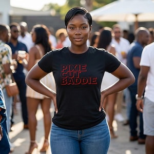 May include: A black short-sleeved t-shirt featuring the red text PIXIE CUT BADDIE printed in a bold, sans-serif font across the chest. The shirt has a crew neckline and a standard fit, paired with blue denim jeans.