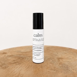 Könnte beinhalten: Eine weiße Rollerflasche für ätherische Ölmischungen mit schwarzem Deckel. Auf dem Etikett steht "calm" in einer minimalistischen Schriftart. Die Flasche steht auf einer Holzoberfläche. Das Produkt hat 10 ml.