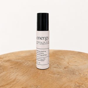Könnte beinhalten: Eine kleine, zylindrische Flasche mit ätherischem Öl-Gemisch, mit schwarzem Deckel und weißem Etikett. Auf dem Etikett steht "energy" und "ESSENTIAL OIL BLEND". Die Flasche steht auf einer Holzoberfläche. Die Flasche enthält 10 ml.