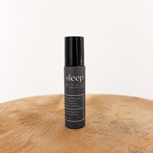 Könnte beinhalten: Eine schwarze Roll-on-Flasche mit ätherischem Öl-Mix mit dem Wort "sleep" in weißer Schrift. Die Flasche hat einen schwarzen Deckel und ein Etikett mit zusätzlichem Text. Die Flasche steht auf einer Holzoberfläche.