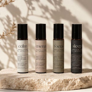 Könnte beinhalten: Vier ätherische Öl-Rollerflaschen mit den Aufschriften "calm", "energy", "focus" und "sleep". Jede Flasche hat einen schwarzen Deckel und ein Etikett mit hellem Text. Die Flaschen werden auf einer hellen, strukturierten Steinoberfläche präsentiert.