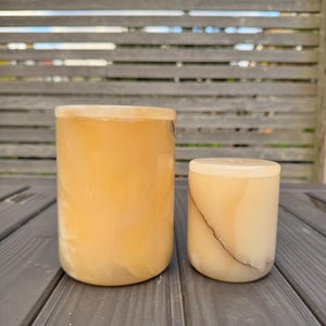 Pode incluir: Dois recipientes cilíndricos de ônix translúcido. O maior é mais alto, o menor é mais curto. Ambos têm uma cor âmbar quente com veios sutis. Eles estão em uma superfície cinza contra um fundo desfocado.