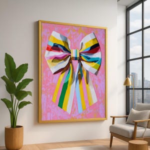 May include: A large framed painting of a colourful bow against a pink background. The bow features stripes of white, yellow, blue, green, and red. The frame is gold. The artwork is displayed in a well-lit room with a plant and a chair.