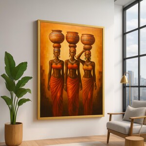 Op de afbeelding: Een ingelijst schilderij toont drie vrouwen in oranje kleding, elk met een terracotta pot op hun hoofd. De vrouwen dragen gouden sieraden en staan tegen een warme, oranje achtergrond. Het kunstwerk is ingelijst in een goudkleurige lijst.