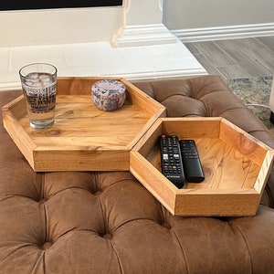 Könnte beinhalten: Zwei sechseckige Holztabletts auf einem braunen, gesteppten Ottomanen. Das obere Tablett hat ein Glas mit Eiswasser und eine kleine dekorative Dose. Das untere Tablett hat zwei schwarze Fernbedienungen.