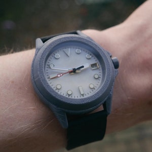 Könnte beinhalten: Eine graue Armbanduhr mit weißem Zifferblatt und schwarzen Zeigern. Die Uhr hat ein Datumsfenster bei 3 Uhr und ein schwarzes Kautschukband.