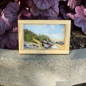 May include: A miniature rectangular painting in a light wood frame. The artwork shows a coastal scene with a sandy path, green grass, rocks, and a blue sky meeting the sea. The painting is set against a backdrop of purple foliage.