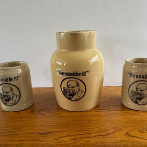 May include: A set of three beige ceramic vessels consisting of one large pitcher and two smaller mugs. Each piece features a black circular graphic of a person holding a glass, with the text "Gesundheit!" printed above the illustration.