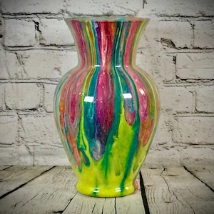 May include: A colorful, hand-painted glass vase with a flared top and base. The vase features vertical streaks of vibrant colors, including pink, green, yellow, and blue. The paint appears to have been dripped down the sides, creating a unique, artistic effect.