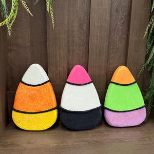 May include: Three candy corn-shaped decorations featuring textured, glittery surfaces. The first is white, orange, and yellow. The second is pink, white, and black. The third is orange, lime green, and purple. Each section is separated by thin black lines.