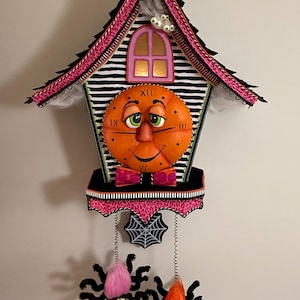 May include: A wall clock shaped like a house with a black and white striped facade, a pink leopard print roof, and a central orange face with green eyes. It features a pink bow tie, a spiderweb pendulum, and two hanging spider ornaments with fuzzy bodies.