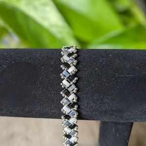 Puede incluir: Un brazalete de cuentas negras y blancas con un patrón de chevron. El brazalete está en un expositor negro.