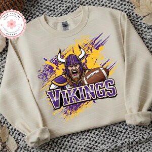 May include: Cream-colored sweatshirt featuring a graphic of a Viking with a football, wearing a purple helmet with horns. The word "VIKINGS" is displayed in purple and white. The design includes yellow and purple paint strokes.