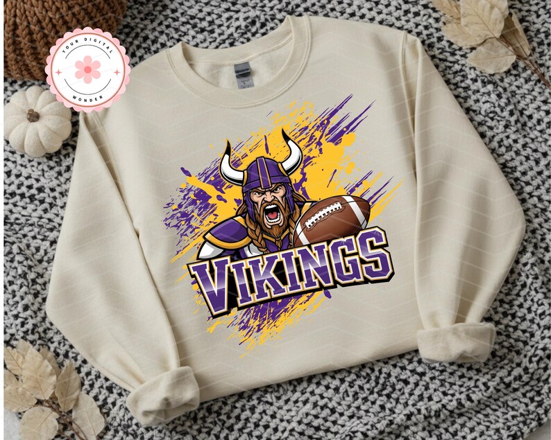 May include: Cream-colored sweatshirt featuring a graphic of a Viking with a football, wearing a purple helmet with horns. The word "VIKINGS" is displayed in purple and white. The design includes yellow and purple paint strokes.