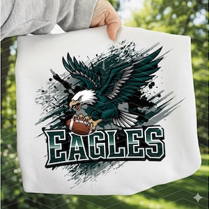 May include: White fabric featuring a detailed graphic of a soaring eagle clutching a football. The eagle is rendered in shades of green, black, and white, set against a backdrop of gray paint splatters. The word "EAGLES" is displayed in bold, green and white lettering.