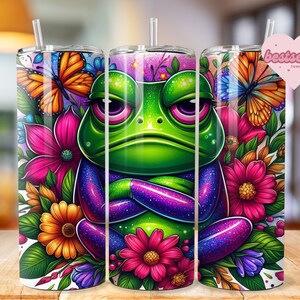 May include: A green frog with purple and blue glitter sitting in a bed of colorful flowers. The frog has its arms crossed and is looking at the viewer. The flowers are pink, yellow, orange, and purple. The background is white.