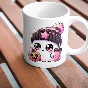 Peut inclure: Mug en c&eacute;ramique blanche avec un fant&ocirc;me de dessin anim&eacute; portant un chapeau ray&eacute; rose et noir, tenant une boisson rose et une petite citrouille orange. Le fant&ocirc;me a de grands yeux et un rougeur rose.