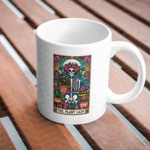May include: White ceramic mug featuring a colourful illustration of a skeleton holding plants. The illustration is framed with a decorative border and the text "The Plant Lady" is at the bottom.