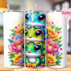 May include: A white tumbler with a design of three cartoon frogs with colorful flowers and leaves. The frogs are stacked on top of each other and have big eyes. The flowers are in shades of pink, yellow, and blue.