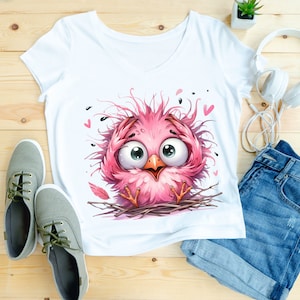 Puede incluir: Camiseta blanca con un p&aacute;jaro rosa de dibujos animados con plumas desordenadas y ojos grandes. El p&aacute;jaro est&aacute; sentado en un nido con algunos corazones rosas en el fondo.