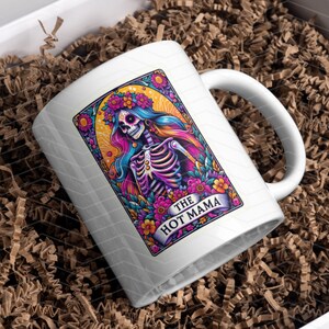 May include: White ceramic mug featuring a colourful illustration of a skeleton with flowers and the text "The Hot Mama".