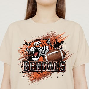 May include: Beige t-shirt featuring a graphic of a roaring tiger and a football. The tiger is orange and black, with the word "BENGALS" in bold, black and white letters below. The design includes orange and black paint splatter.