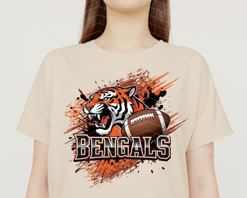 May include: Beige t-shirt featuring a graphic of a roaring tiger and a football. The tiger is orange and black, with the word "BENGALS" in bold, black and white letters below. The design includes orange and black paint splatter.