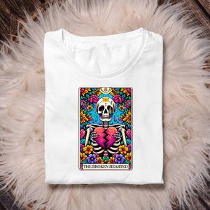May include: White t-shirt featuring a colourful illustration of a skeleton holding a broken heart. The illustration is surrounded by flowers and the text "The Broken Hearted".