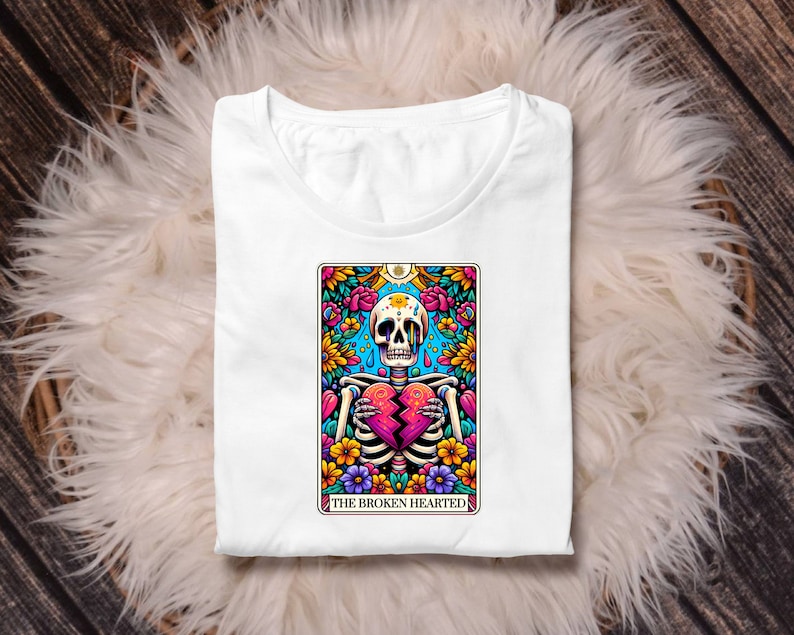 May include: White t-shirt featuring a colourful illustration of a skeleton holding a broken heart. The illustration is surrounded by flowers and the text "The Broken Hearted".