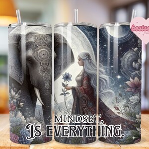 Può includere: Un bicchiere bianco con un design colorato che presenta una donna con lunghi capelli bianchi, un elefante e fiori. Il testo "MINDSET IS EVERYTHING." è stampato sul bicchiere.