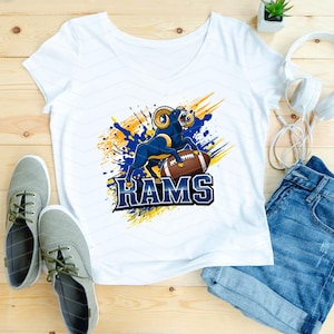 May include: White v-neck t-shirt featuring a blue and gold graphic of a ram with a football and the word "RAMS". The shirt is paired with denim shorts, gray sneakers, and white headphones.