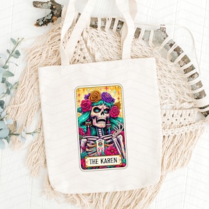 May include: A white tote bag featuring a colourful illustration of a skeleton wearing a flower crown. The illustration is in the style of a tarot card and says "The Karen" at the bottom.