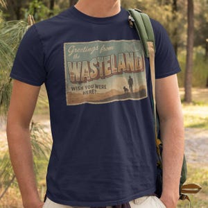 以下が含まれることがあります： ヴィンテージポストカードのデザインが施されたネイビーブルーのTシャツ。「Greetings from the Wasteland, Wish You Were Here?」と書かれたポストカードには、人物と犬のシルエットが描かれています。Tシャツはクルーネックで半袖です。
