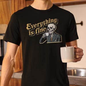 Może przedstawiać: Czarny T-shirt z nadrukiem szkieletu w garniturze trzymającego filiżankę kawy. Na koszulce widnieje napis "Everything is fine" w kolorze żółtym.