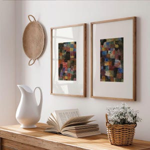 May include: Two framed rectangular wall art pieces featuring colorful patchwork patterns, a white ceramic pitcher, an open book with visible text, and a small woven basket filled with white flowers arranged on a wooden surface.