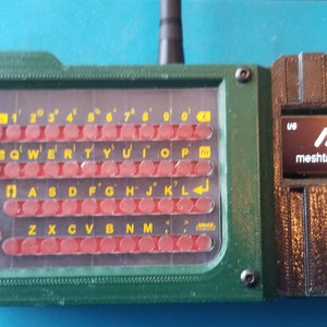May include: A green and black 3D printed device with a keyboard and a small screen displaying "mesh.tastics.org". The screen is labeled "UV" and "3.3V".