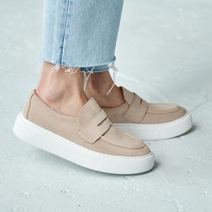 May include: Beige suede loafers with a white sole. The shoes, with a classic penny loafer design, are worn with light blue denim jeans. The jeans have a frayed hem.