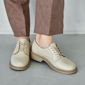 May include: Cream-colored leather oxford shoes with a rounded toe and a thick, light brown sole. The shoes are laced up and paired with brown trousers. The image is taken from a low angle, showing the shoes and the lower part of the legs.