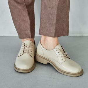 Beige leather derby shoes for women featuring a minimalist lace-up design with round toes and sturdy low block heels. Handmade from genuine leather, these versatile oxford-style shoes offer comfort and durability, perfect for casual wear, office outfits, or smart-casual looks. A timeless neutral tone makes them easy to pair with trousers, jeans, or dresses, adding a modern yet classic touch to everyday fashion.