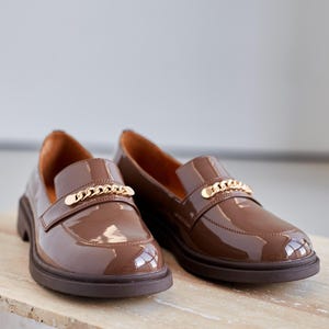 Women’s brown patent leather loafers with a glossy finish and chunky sole. Designed with a gold chain detail across the upper for a modern, stylish look. Slip-on style makes them versatile and comfortable for everyday wear, office outfits, or casual chic streetwear. Durable sole provides support while the polished surface adds elegance, making these loafers a perfect blend of sophistication and practicality for fashion-forward women.