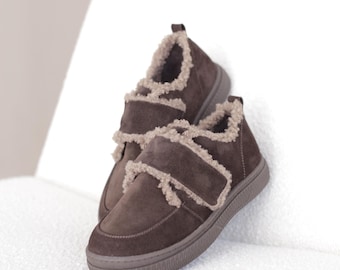 Op de afbeelding: Een paar donkerbruine suède sneakers met een klittenbandsluiting over de wreef. De schoenen zijn afgewerkt met een lichtbeige imitatiebontrand langs de kraag en de randen van de bandjes, geplaatst op een bijpassende donkerbruine rubberen zool.