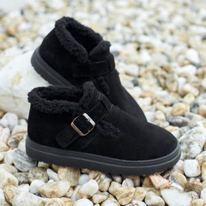 May include: A pair of black suede ankle boots with a buckle closure and faux fur lining. The boots have a round toe and a thick, black sole. The shoes are displayed on a bed of small, light-colored stones.