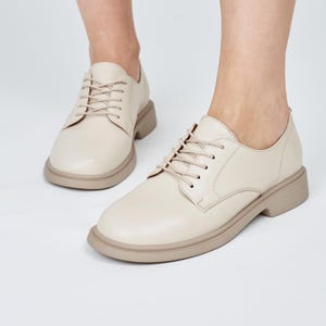 May include: Cream-colored derby shoes with a rounded toe and a thick, light brown sole. The shoes have a lace-up closure with matching cream-colored laces. The shoes are displayed on a light background, showcasing their design and color.