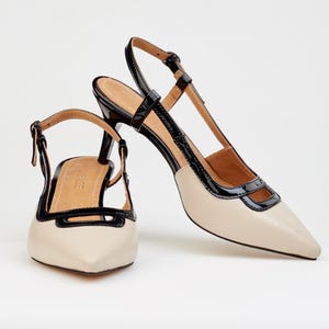 May include: Pair of cream-colored slingback heels with black patent leather trim. The shoes have pointed toes and a thin strap that buckles around the ankle. The heels are approximately 3 inches high.