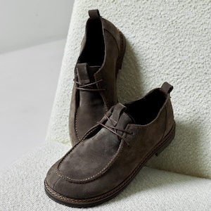 May include: A pair of dark brown suede shoes featuring a moccasin-style toe, thin brown laces, a pull tab at the heel, and a dark brown sole with visible stitching along the edge. The shoes have a low-top silhouette and a soft, matte texture.