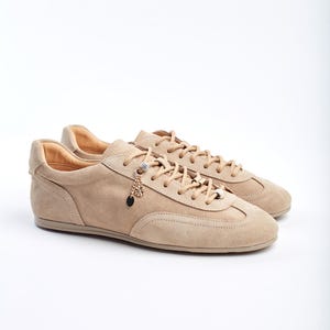 Low-top women’s sneakers in soft beige suede, with a streamlined silhouette and tonal laces threaded through metal eyelets and decorative charm accents. The supple suede upper features subtle stitching lines and curved paneling for a retro-inspired yet modern look. A cushioned leather-lined interior and slim rubber sole provide all-day comfort and traction, making these versatile trainers ideal for casual weekend wear or stylish everyday outfits.